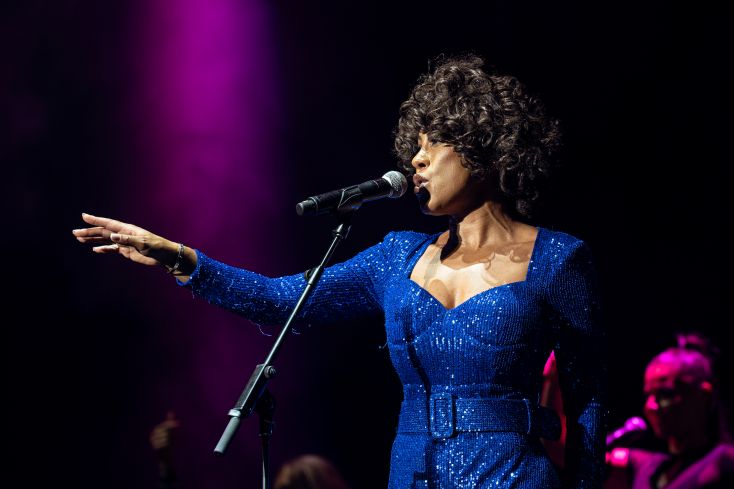 A singer styled as Whitney Houston stands with her hand outstretched
