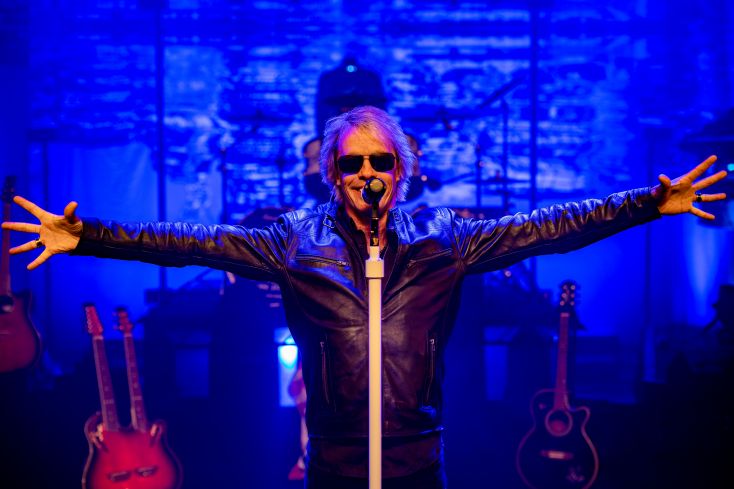 A tribute artist to Bon Jovi stands with his arms outstretched wide.