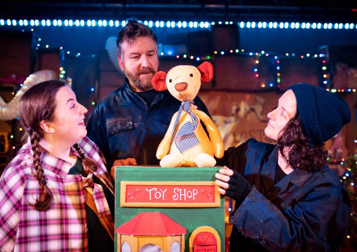 One actor and two actresses stand around a teddy bear, looking at it as if listening to it tell a story.