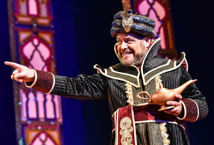 John Thompson dressed in an ornate costume holds Aladdin's lamp and points menacingly.