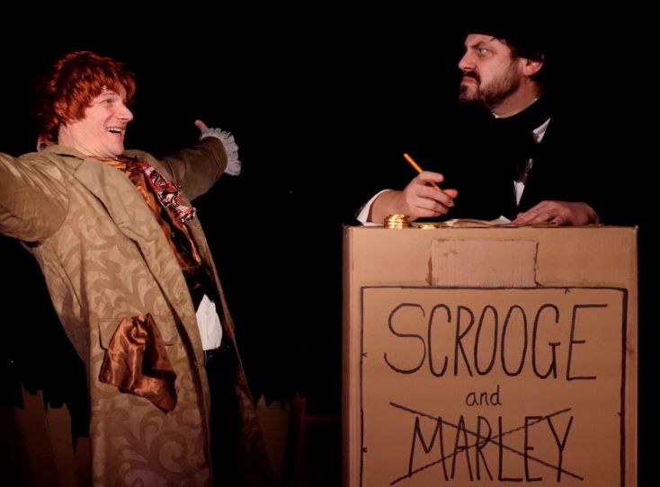 Two actors stand facing each other, one with his arms out wide and joyful, the other scowls at this behaviour.