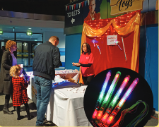 A man, a woman and a child stand in front of a table. A lady is standing behind the table selling merchandise. Behind her is a backdrop that looks like curtains. Inset image of bright light wands.