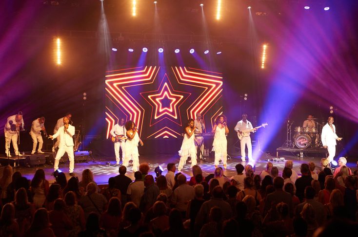 A stage with a group of four performers singing accompanied by musicians. All are dressed in white with backdrop of a star.